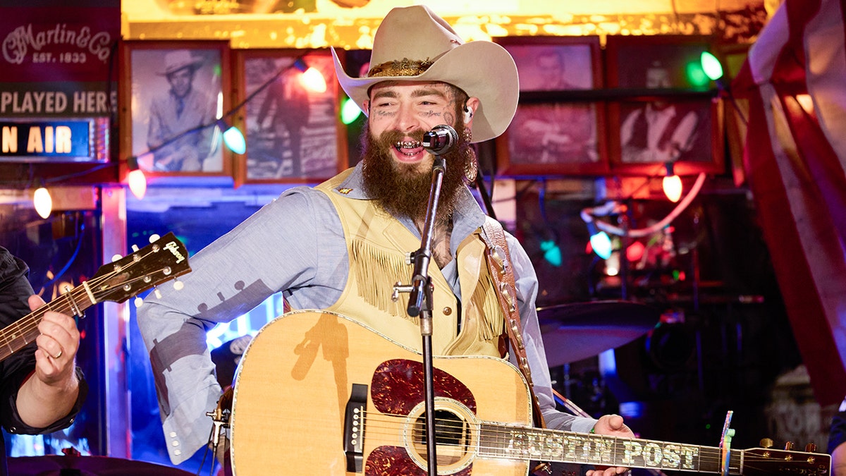 Post Malone smiling on stage wearing a tan cowboy hat, yellow vest, and blue shirt