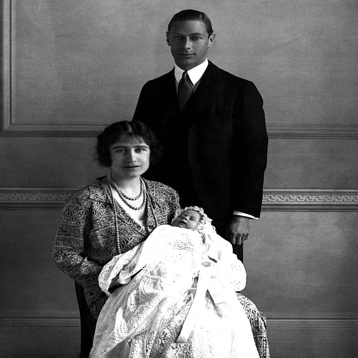 A formal vintage family portrait with a seated woman holding a baby in christening attire, and a man standing behind them in a suit