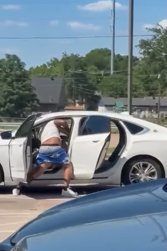 A man in a white tank top and blue shorts appears to be entering or exiting the driver's side of a white sedan with all doors open.