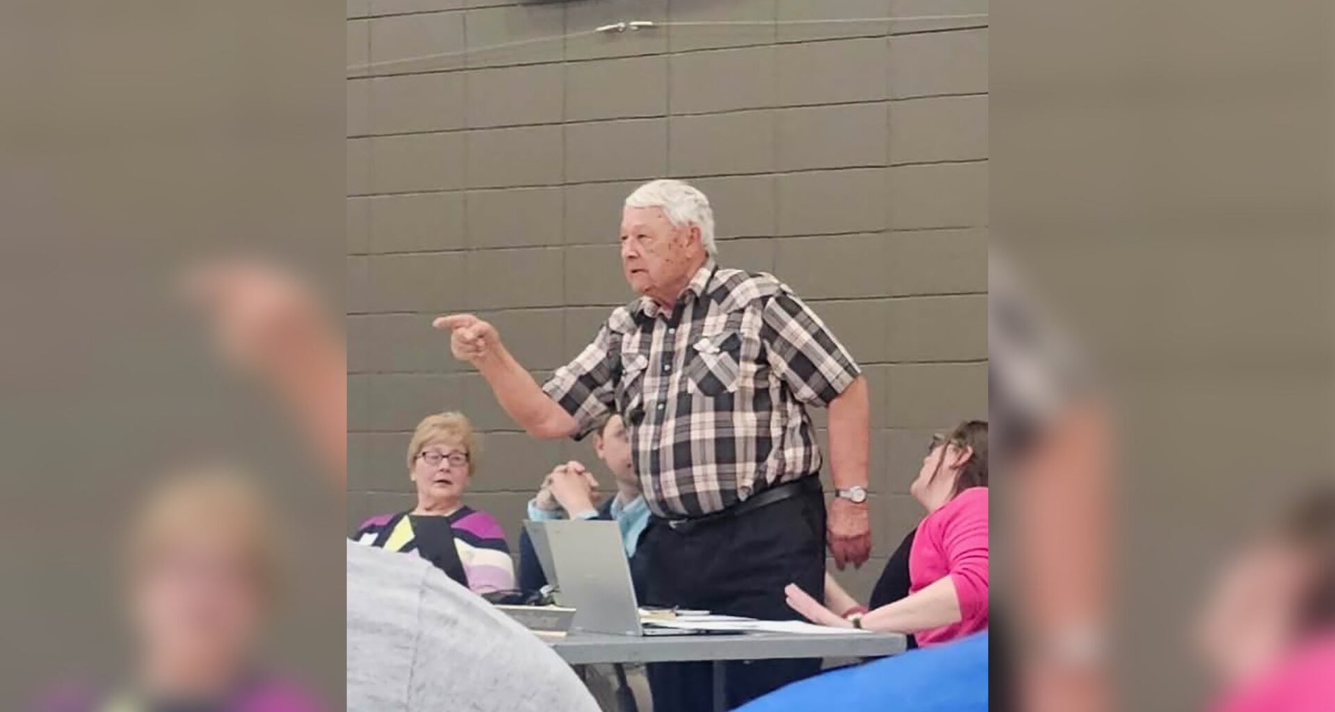 An older man in a plaid shirt stands and points while speaking at a table with seated people in a room with gray cinder block walls.