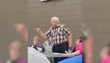 An older man in a plaid shirt stands and points while speaking at a table with seated people in a room with gray cinder block walls.