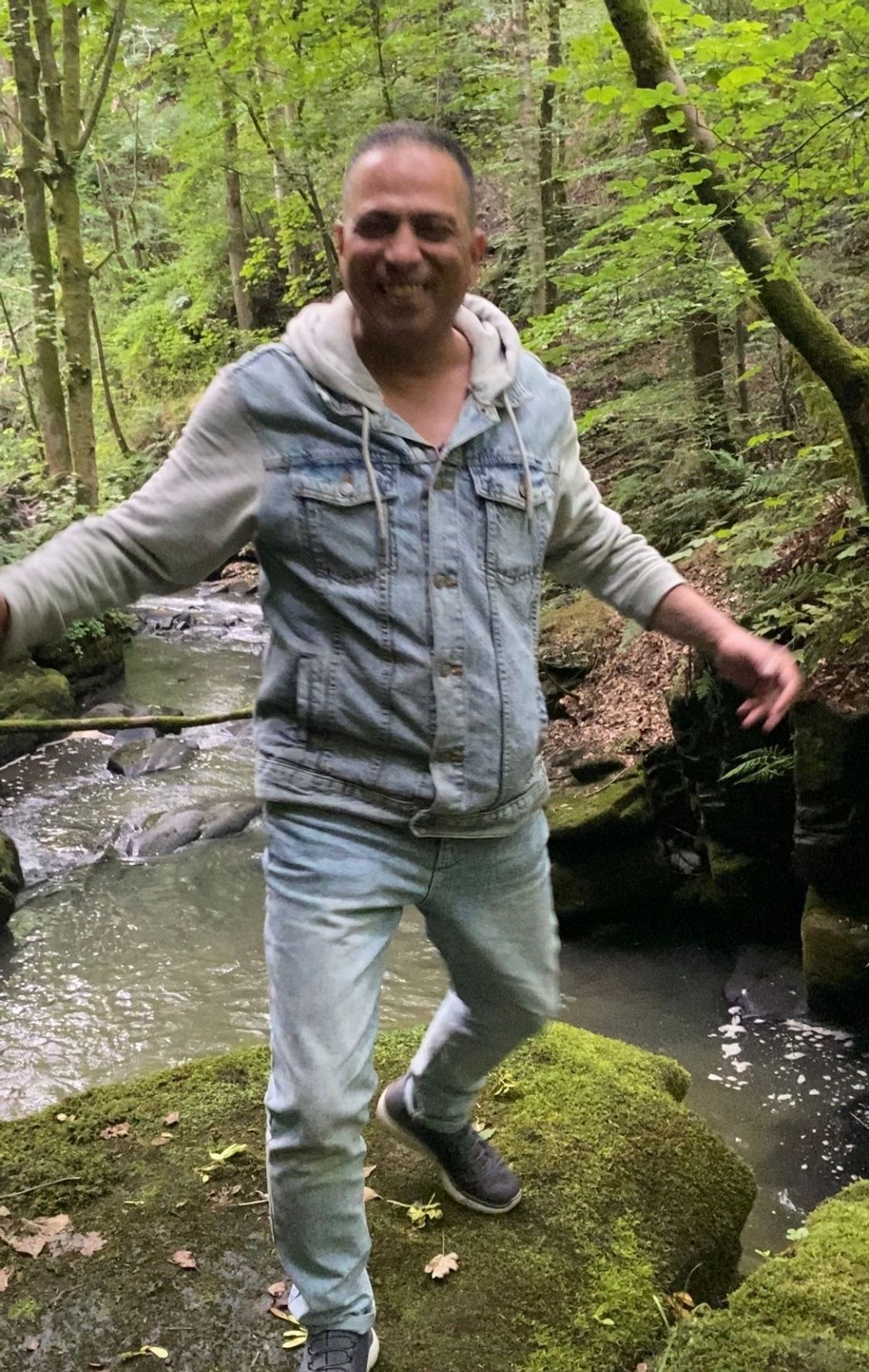 Mohamed smiling as he stands on rocks in a strem wearing jeans and a denim jacket