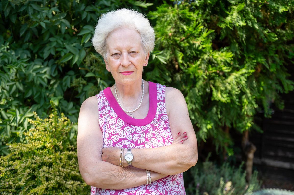 An older woman with white hair stands confidently with her arms crossed, positioned in a lush garden with vibrant green foliage in the background. She wears a sleeveless floral top and a pink necklace, radiating a sense of poise and tranquility.