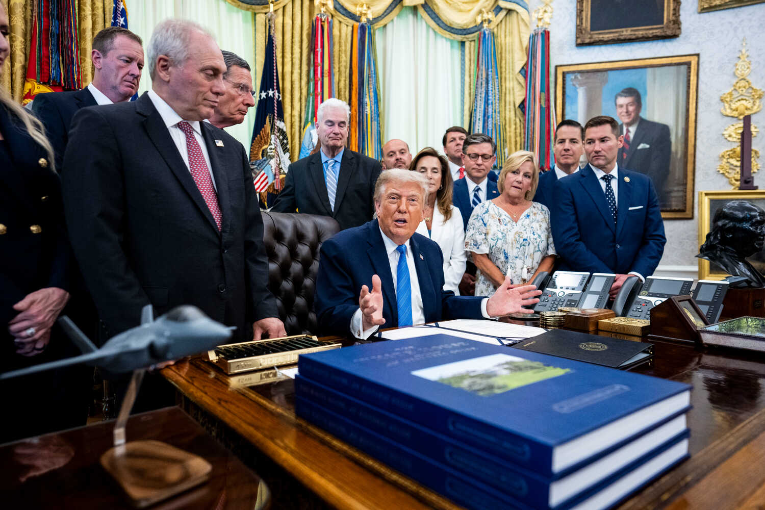 President Trump, wearing a navy suit, white shirt and blue tie, gestures as he sits at a desk. Standing behind him are about a dozen people.