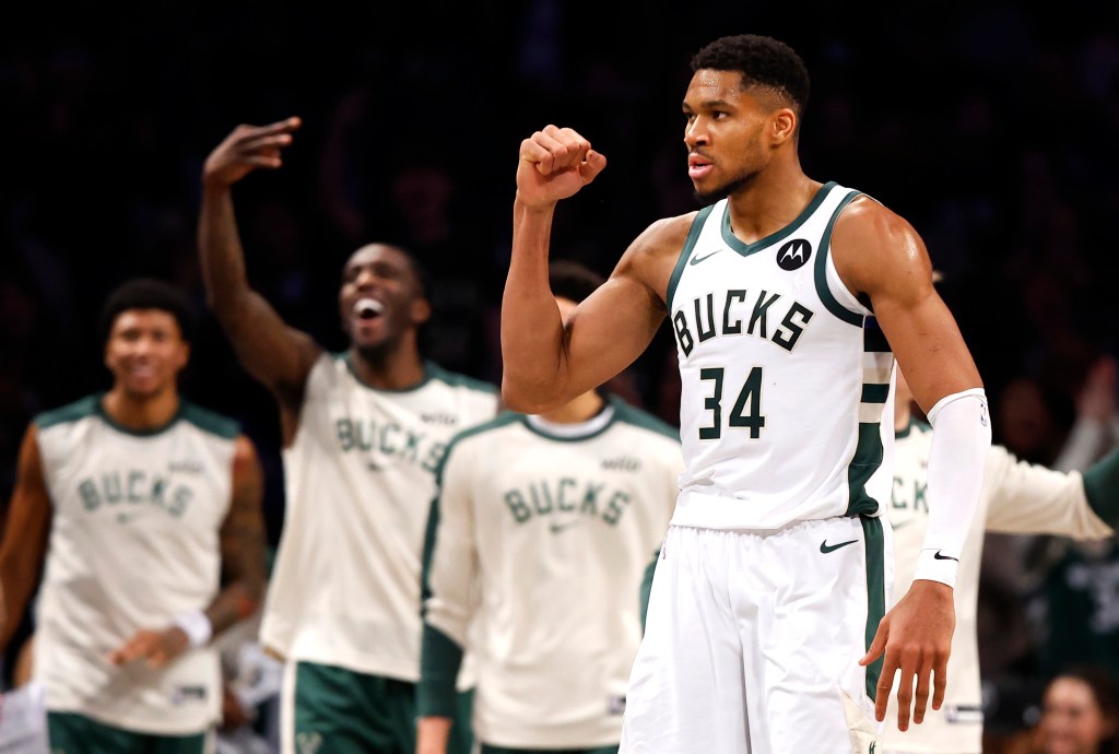 Giannis Antetokounmpo in a white Bucks jersey with number 34, flexing his arm with other players in the background.
