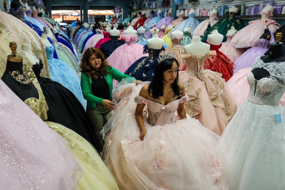 A woman straightens the skirt of a girl wearing a tiara and a formal gown with full skirt amid mannequins in gowns.
