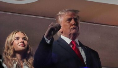 Donald Trump addressing media at a press event, wearing a suit and tie, with reporters and cameras in the background.