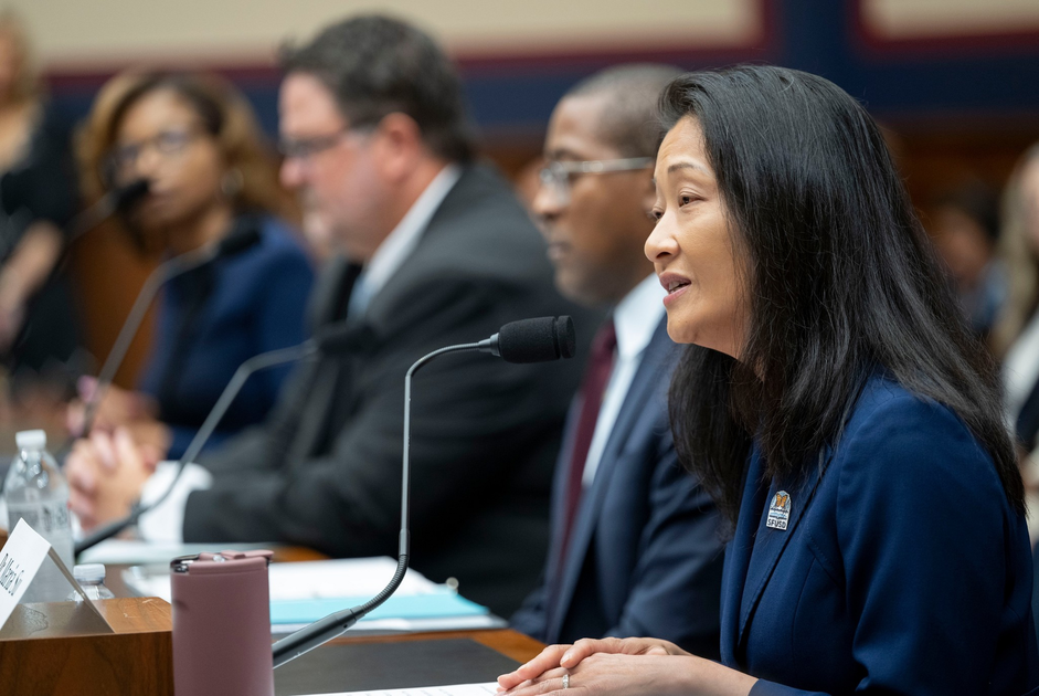 Four people in formal attire sit at a panel with microphones, with the woman in the foreground speaking during a formal meeting or hearing.