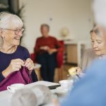 Senior female friends spending leisure time knitting while sitting at table in retirement home - stock photo