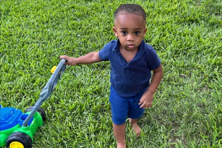 Kohen Wiley in a blue outfit stands outside in the grass, one hand on a toy lawnmower