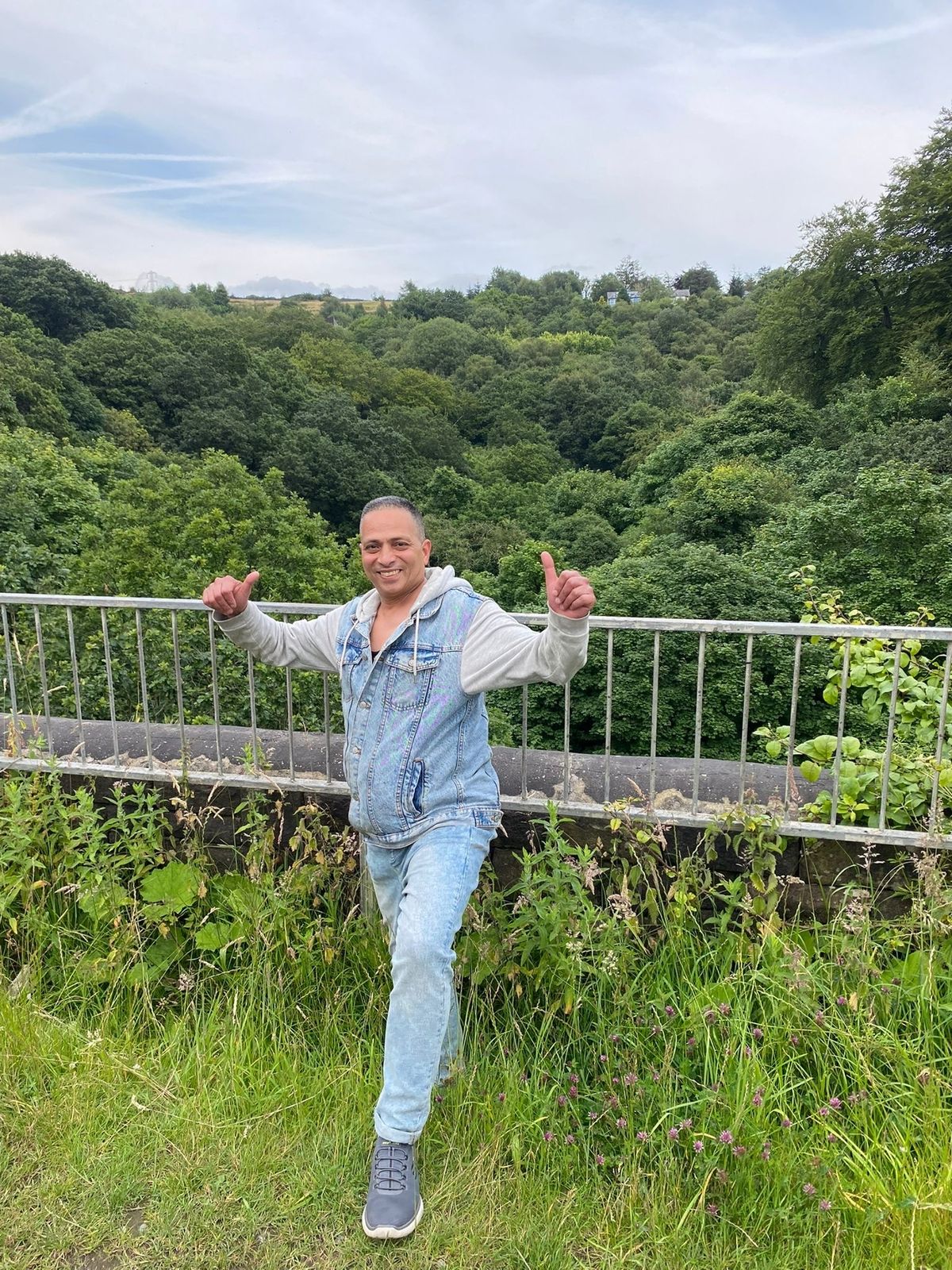 Mohamed stands with both thumbs up against a metal fence surrounded by trees