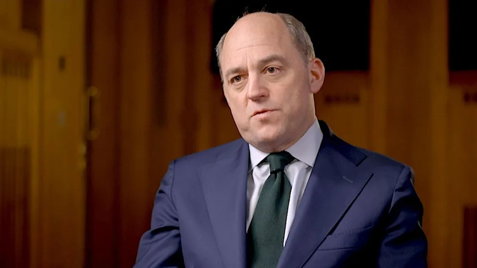 Ben Wallace, a greying hair at the sides of his head, wearing a navy suit and a dark tie, sitting in a dark, wood-panelled room
