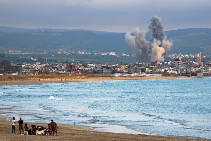 A coastal view from a sandy beach where several people are gathered, looking across the water toward a seaside town where a massive plume of gray and white smoke rises from an explosion.