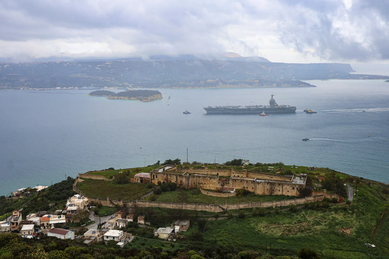 An elevated view of a coastal landscape featuring an old stone fortress on a grassy hill overlooking a large body of water. A large military aircraft carrier sails in the distance under a cloudy sky.