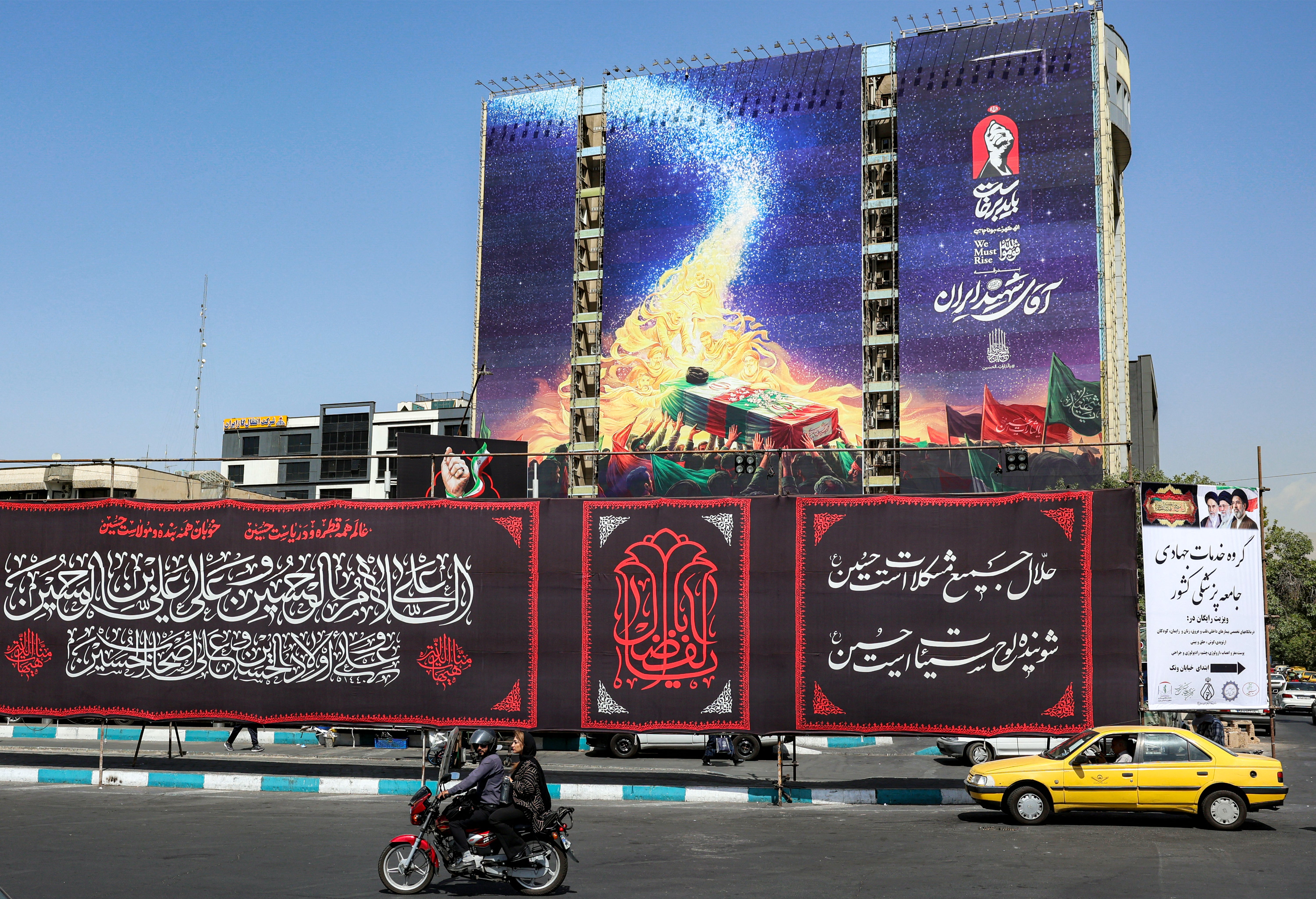 Motorists drive vehicles past a mural displayed on the facade of a building depicting late figures aligned with the Iranian government, such as late President Ebrahim Raisi, slain Islamic revolutionary guards commander Qasem Soleimani, slain Lebanese Hezbollah leader Hassan Nasrallah, and others