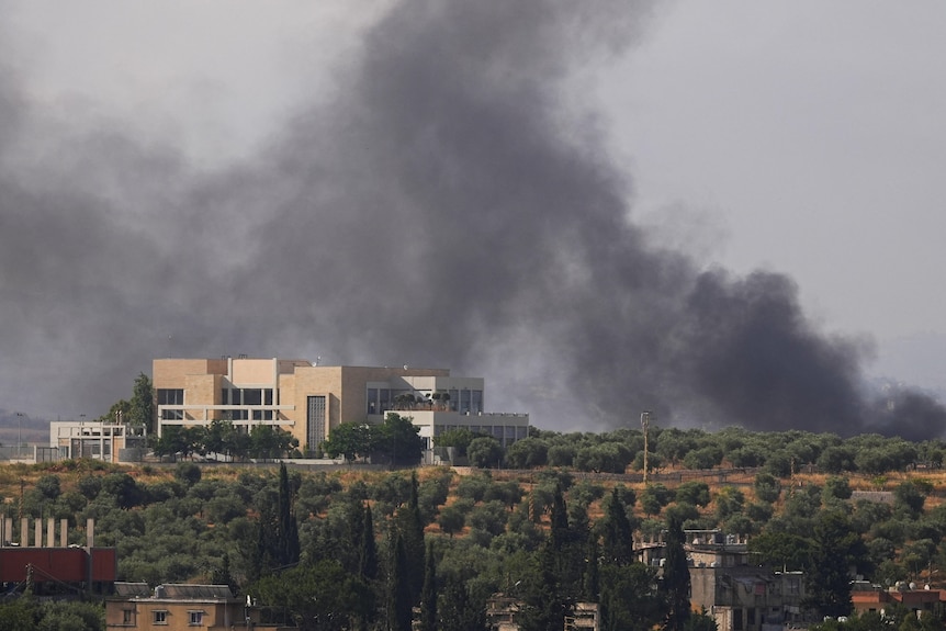 A building stands on the top of a hillside, with grey smoke rising behind it into the sky.