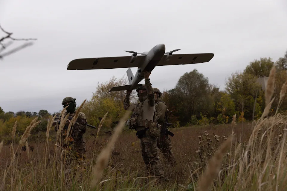 Figures in camouflage in a brown field launch a grey drone in front of trees and under a grey sky
