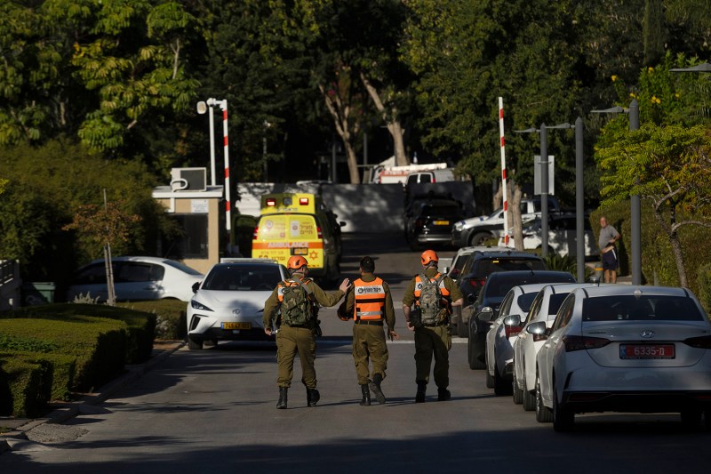 Three emergency personnel in green and orange uniforms walk away from the camera down a tree-lined street parked with cars, toward a yellow ambulance in the background.