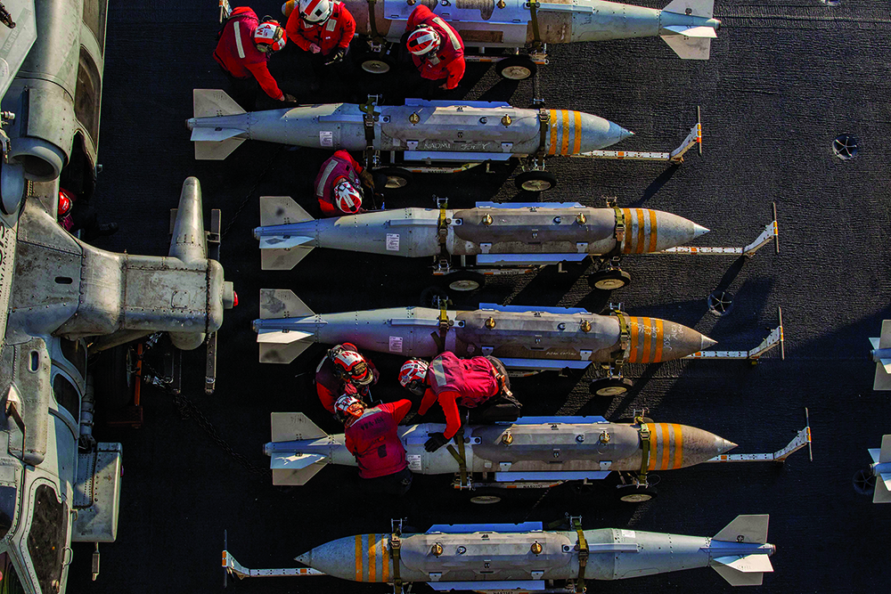 An overhead view of a military flight deck where crew members in red uniforms work alongside rows of large, gray bombs arranged horizontally next to a parked aircraft.
