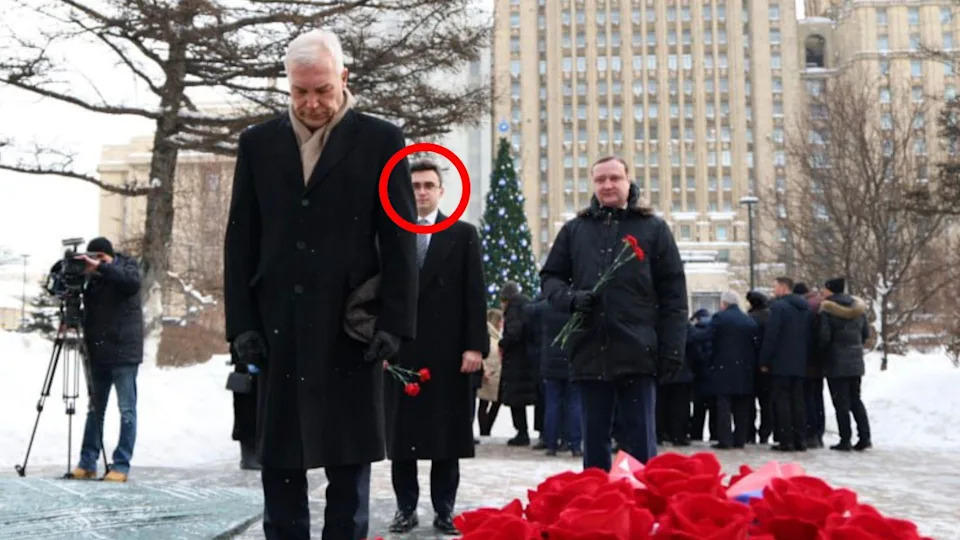 Russian Deputy Foreign Minister Alexander Grushko, a man with white hair, wearing a dark overcoat and a scarf, stands with his head bowed, in front of a display of red flowers seemingly laid as a memorial. Behind him stands Evgeny Lyukshin, a young man with dark hair and glasses, wearing a dark overcoat and tie, holding a couple of red flowers. There is a crowd of people further behind in what appears to be a snowy Moscow public square them and a man is filming the event with a camera on a tripod.