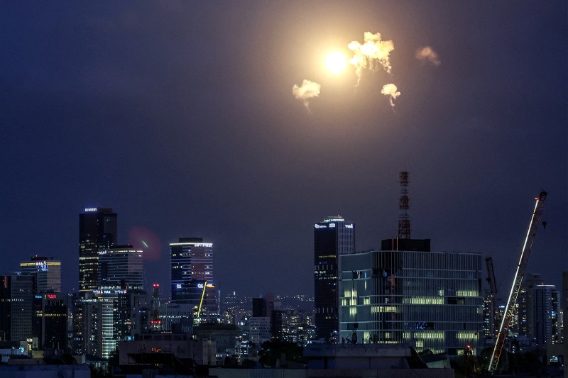 A night cityscape featuring several lit skyscrapers under a dark sky, with a bright flash of light and small clouds of smoke visible in the upper atmosphere.