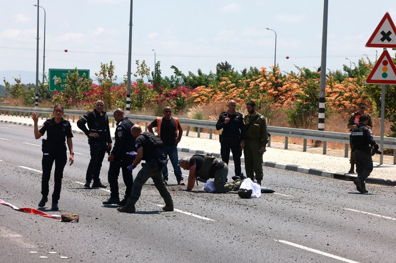 A group of uniformed emergency and security personnel gather on an asphalt road scattered with debris, with one individual kneeling to inspect an item on the ground.