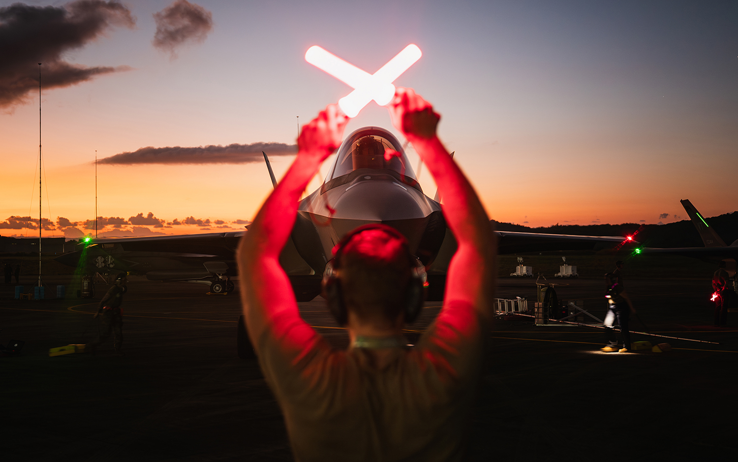 A ground crew member, seen from behind, raises two glowing red light sticks in an "X" position to guide a military fighter jet on a runway during a vibrant orange sunset.
