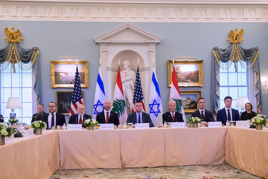 Men and women sit around a long table with multiple flags behind them.