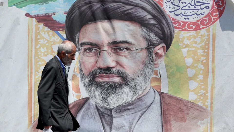 A man with grey hair wearing a dark suit and blue shirt carrying a plastic bag walks past a poster of Supreme Leader Mojtaba Khamenei who has a grey beard and is wearing a grey buttoned shirt, maroon shawl glasses and a black turban.