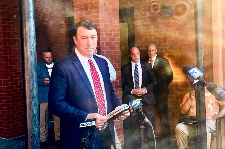 A man in a navy suit and red tie speaks at a press conference outside of a red brick building