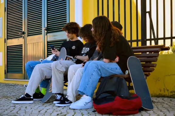 A group of teenagers sit on a bench with skateboards, looking at their phones and talking outside a building.