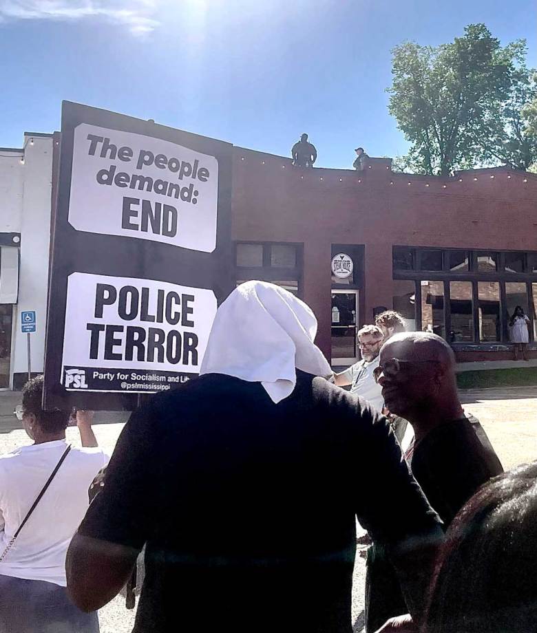 People gather on the street, one holding a sign that reads "The people demand: END POLICE TERROR. Police can be seen on the rooftop across the road