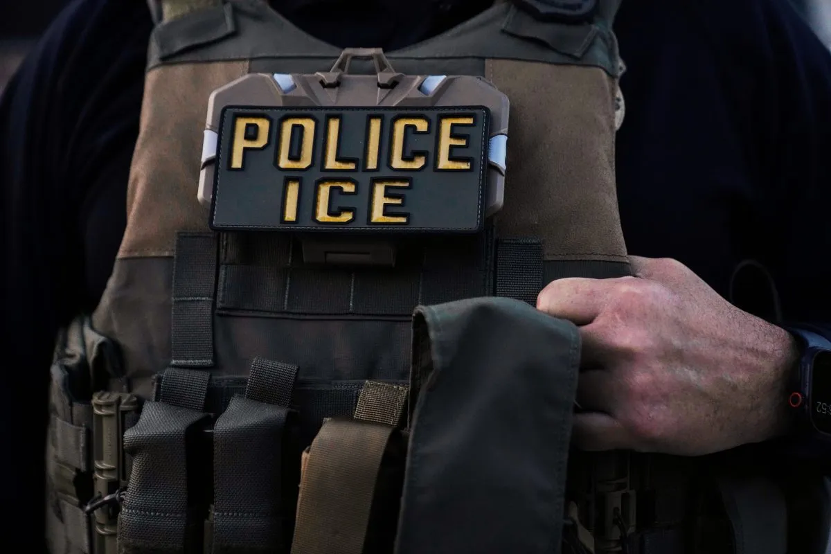 A person wearing a tactical vest with a prominent patch reading "POLICE ICE," often associated with ICE detentions, holds the front of the vest with one hand.