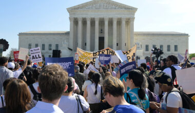 Protestors stand outside the Supreme Court with signs on a sunny day.
