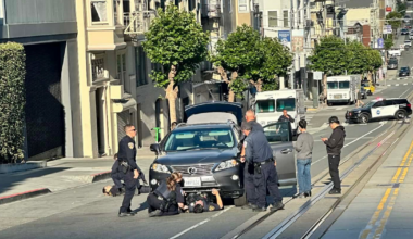 Police officers have detained a person lying face-up on the street in front of a gray Lexus with its trunk open, while two bystanders stand nearby.