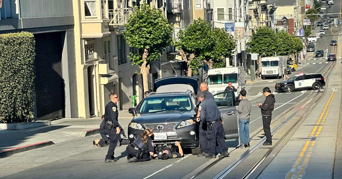 Police officers have detained a person lying face-up on the street in front of a gray Lexus with its trunk open, while two bystanders stand nearby.