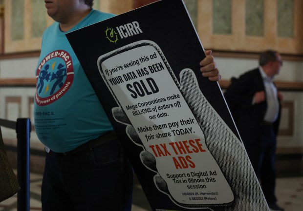 A demonstrator holds a sign referencing corporations that make money through the sale of peoples' personal data before a rally held by several groups under the umbrella organization Illinois Revenue Alliance at the State Capitol during the spring legislative session on May 27, 2026, in Springfield. (John J. Kim/Chicago Tribune)