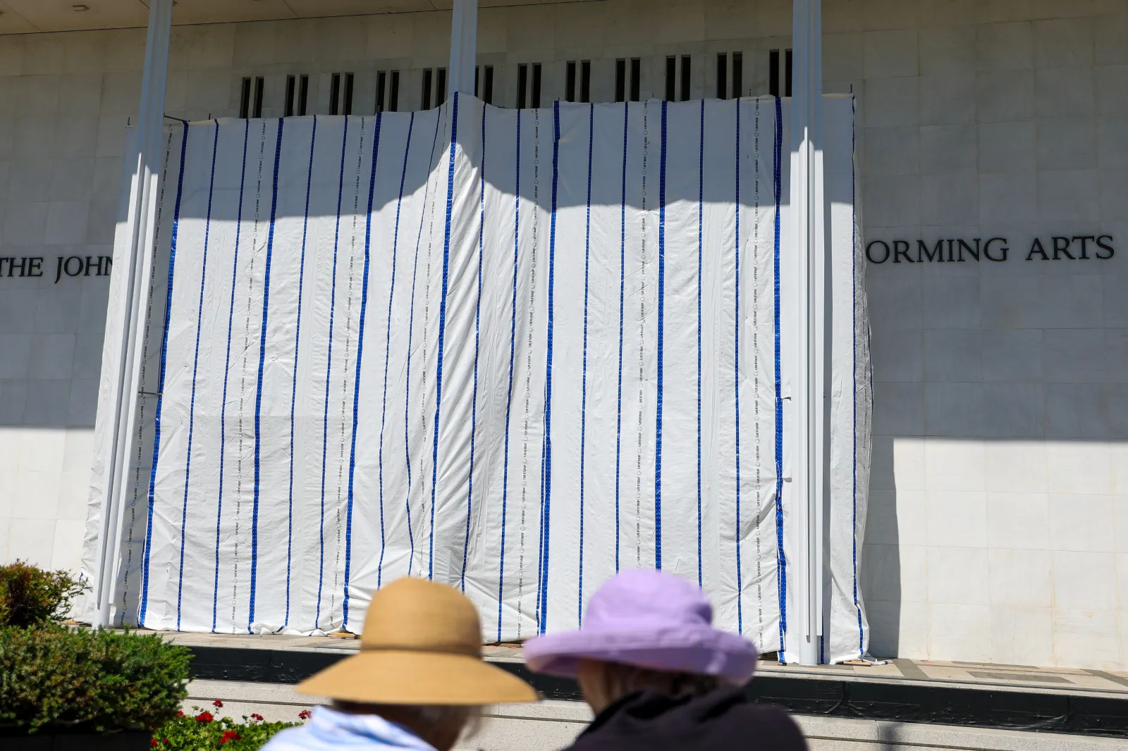 People gather to watch as tarps are hung on scaffolding outside The Kennedy Center on June 13, 2026 in Washington, DC. Friday late night workers removed the President Donald Trump's name from the Kennedy Center.