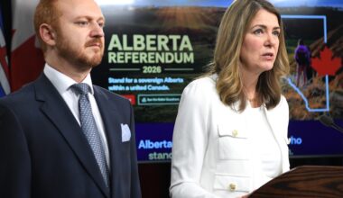 A 44-year-old man with reddish hair and wearing a blue suit stands beside Danielle Smith, who is wearing a white suit. Behind them is a monitor that says &lsquo;Alberta Referendum 2026.&rsquo;