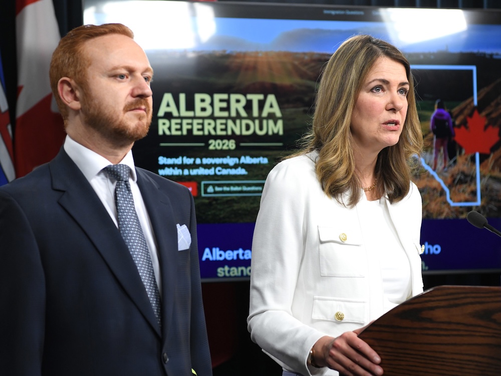 A 44-year-old man with reddish hair and wearing a blue suit stands beside Danielle Smith, who is wearing a white suit. Behind them is a monitor that says &lsquo;Alberta Referendum 2026.&rsquo;