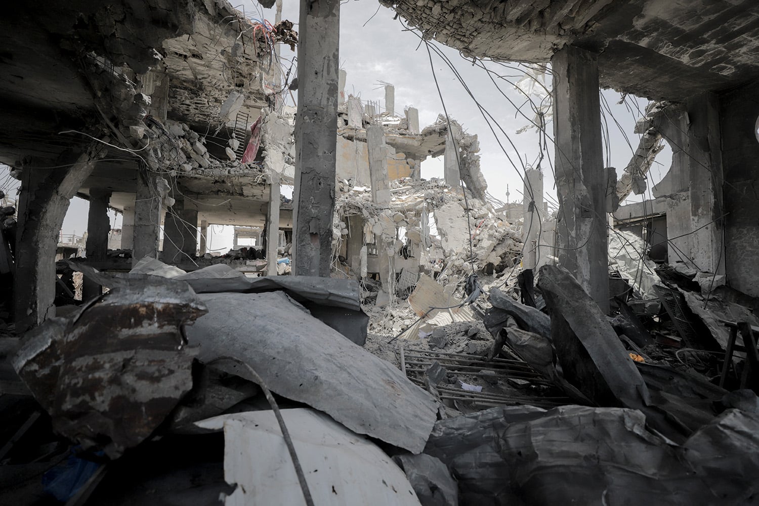 A wide shot from inside a heavily destroyed concrete building, looking out through large gaps where walls used to be. The foreground is filled with mounds of rubble, twisted metal, and shattered debris. Exposed concrete pillars and sagging, broken ceilings frame the view of another multi-story building in the background that has completely collapsed into a pile of white dust and concrete fragments. Exposed rebar hangs loosely from the structures under an overcast sky.