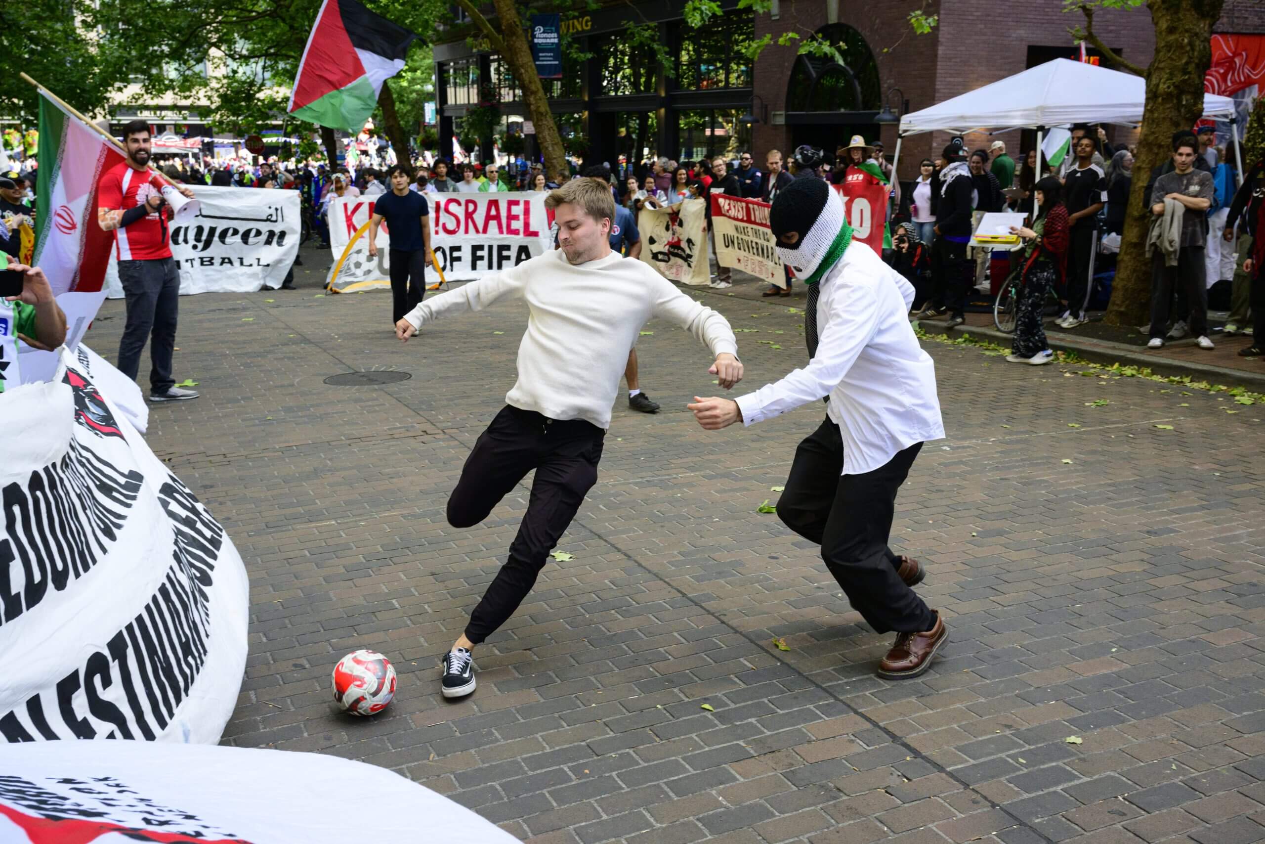 Two people kick a football in a square surrounded by protests