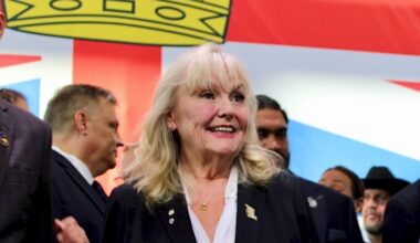 A woman in her 70s with shoulder-length blond hair smiles as a crowd surrounds her. A large British Columbia flag can be seen behind her.