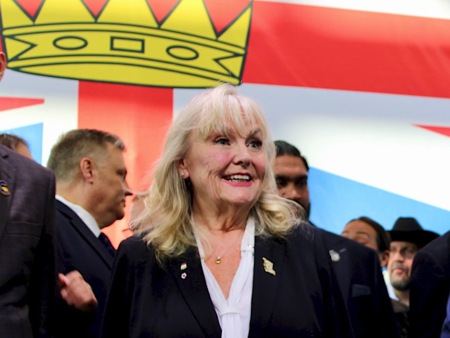 A woman in her 70s with shoulder-length blond hair smiles as a crowd surrounds her. A large British Columbia flag can be seen behind her.