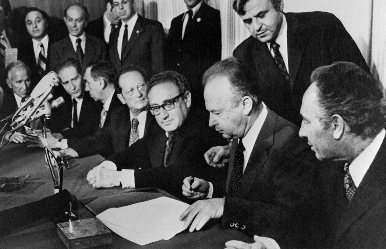 A black and white photograph of a group of men gathered around a long table. In the center, a man in a dark suit leans forward to sign a document with a pen, while a man wearing glasses directly to his left watches him closely. Several other men in formal suits stand and sit around them, looking toward the table or the signing process. Multiple microphones are clustered together on the left side of the table.