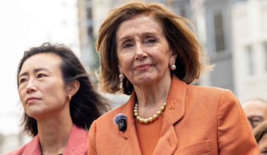 Two women stand outdoors wearing orange blazers; the woman on the right has pearl earrings and a pearl necklace, looking directly ahead with a serious expression.