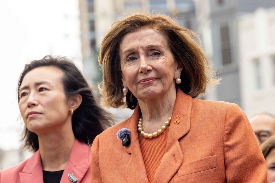 Two women stand outdoors wearing orange blazers; the woman on the right has pearl earrings and a pearl necklace, looking directly ahead with a serious expression.