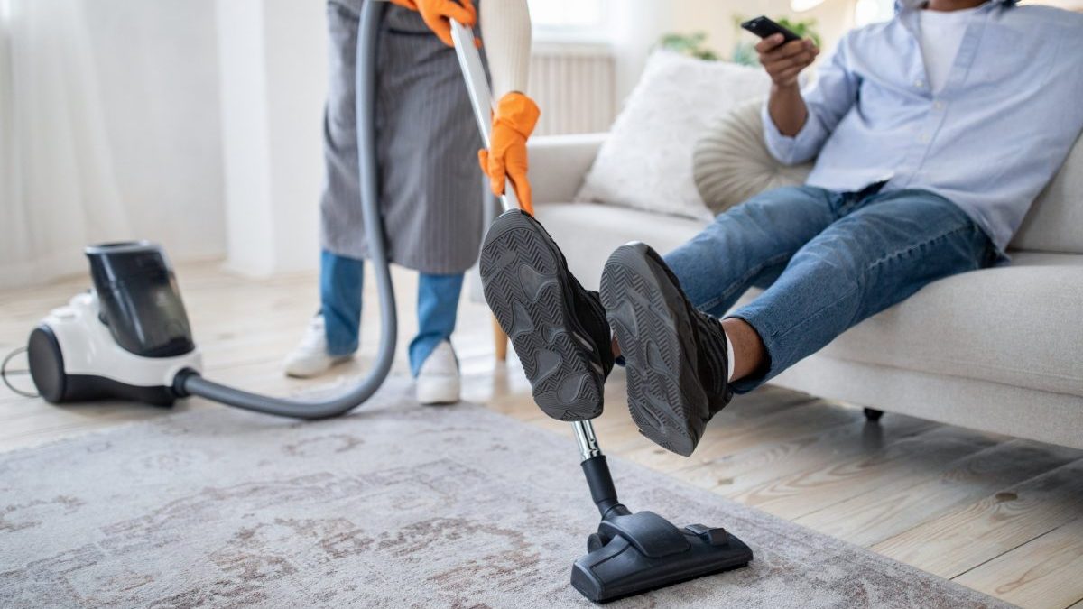 Millennial black lady vacuuming floor while her husband watching TV, not helping her with household duties, cropped view. Unrecognizable young couple having problem with domestic chores