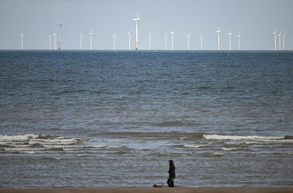 The Ryhl Flats windfarm is pictured on the horizon in the Irish Sea as a dog walker strolls on the beach at Abergele in north Wales on April 21, 2026. Britain's government on April 21, 2026, unveiled plans to accelerate its clean energy drive after oil and gas prices soared in the wake of the US-Iran war. Placing solar panels and wind turbines on industrial and railway sites could power about five million homes, the Labour government said in a statement. (Photo by Paul ELLIS / AFP via Getty Images)