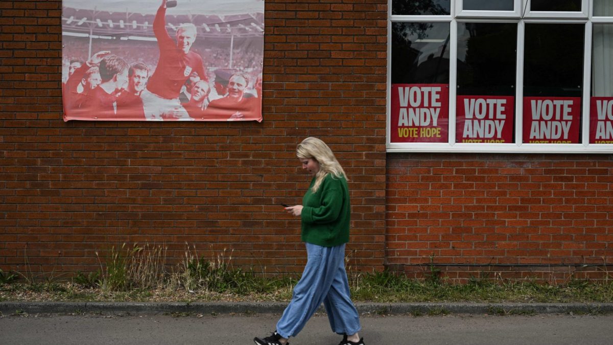 A woman walks past campaign posters (R) for the Labour party candidate, Greater Manchester Mayor Andy Burnham, and an old poster of England football player Bobby Moore (L) hoisting the 1966 World Cup trophy, along a street ahead of the Makerfield by-election on June 18, west of Manchester, north-west England on June 16, 2026. Britain holds a special vote on June 18 that is expected to set in motion an attempt to oust Labour leader Keir Starmer as prime minister. (Photo by Oli SCARFF / AFP via Getty Images)